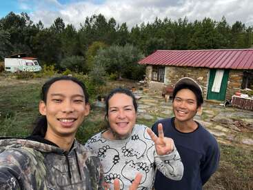 Tres personas sonríen y hacen signos de paz al aire libre cerca de una casa rústica de piedra con tejado rojo. Al fondo, frondosos árboles verdes y una autocaravana.