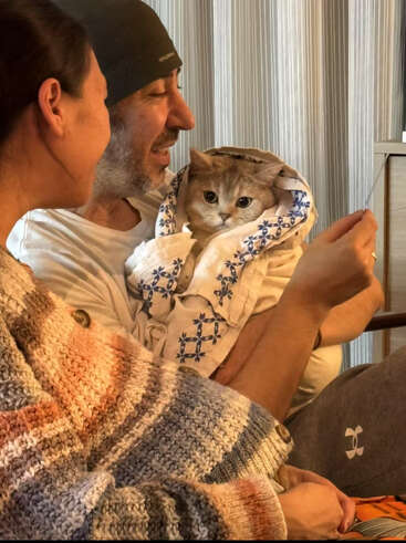 Ein Mann und eine Frau lächeln zusammen und halten eine Katze, die in ein dekoratives Handtuch eingewickelt ist. Die Katze sieht gemütlich und ruhig aus und schafft eine warme, fröhliche Atmosphäre.