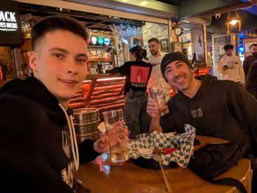 Zwei Freunde sitzen an einer Bar, lächeln und halten Biergläser in der Hand. Die Atmosphäre ist lebhaft, mit Menschen und Barkeepern im Hintergrund. Ein leerer Essenskorb ist zu sehen.