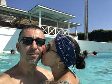Ein Paar genießt einen sonnigen Tag in einem Pool. Die Frau küsst den Mann auf die Wange. Beide tragen eine Sonnenbrille; im Hintergrund ist ein weißes Gebäude zu sehen.