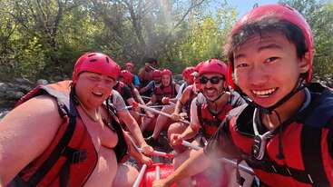 Eine Gruppe von Menschen, die alle rote Helme und Schwimmwesten tragen, posiert fröhlich beim Wildwasser-Rafting an einem sonnigen Tag umgeben von grünen Bäumen.