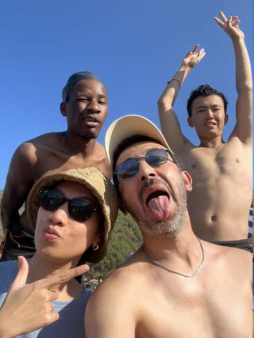 Vier Freunde posieren freudig für ein sonniges Strand-Selfie. Einer streckt die Zunge heraus, die anderen machen spielerische Gesten, alle lächeln bei strahlend blauem Himmel und Sonnenlicht.