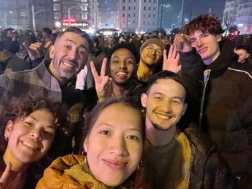 Eine Gruppe von sechs Freunden lächelt fröhlich für ein nächtliches Selfie, umgeben von einer großen Menschenmenge in einer hell erleuchteten städtischen Umgebung.