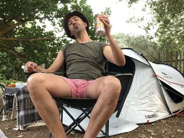 A man relaxes in a camping chair, wearing a hat and red shorts, holding a drink, with a tent and trees in the background. Outdoor setting.