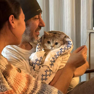 Um homem e uma mulher sorriem juntos, segurando um gato enrolado em uma toalha decorativa. O gato parece aconchegante e calmo, criando uma atmosfera calorosa e alegre.