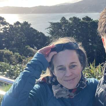 Uma mulher com uma jaqueta azul sorri, segurando óculos escuros na cabeça. Atrás dela, árvores e água brilham à luz do sol, com colinas à distância.