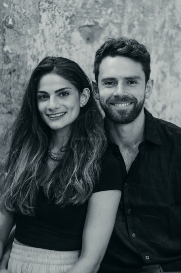 Das Bild ist ein Schwarz-Weiß-Foto von einem Mann und einer Frau, die zusammen posieren, wobei die Frau links und der Mann rechts steht und beide in die Kamera lächeln.