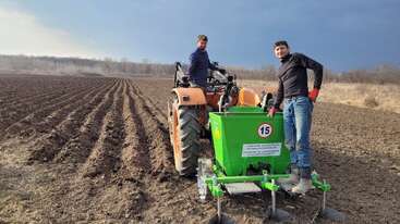 Two men are working with a tractor and green farming equipment in a freshly plowed field. The sky is cloudy, and trees border the agricultural area.