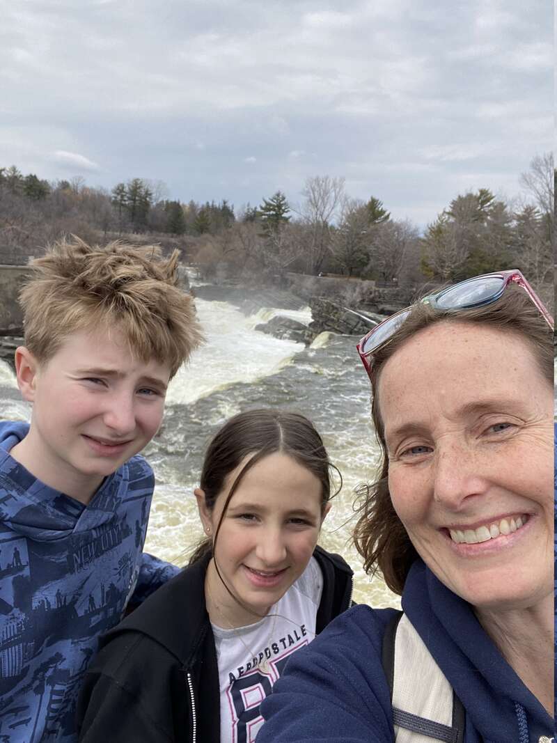 Una mujer y dos niños sonríen para una selfie frente a una cascada, con un cielo nublado y árboles sin hojas como pintoresco telón de fondo.