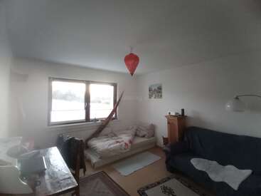 Esse quarto aconchegante tem um sofá-cama, sofá azul escuro, escrivaninha de madeira, janela grande, lanterna vermelha, tapetes e decoração minimalista, criando uma atmosfera simples e convidativa.