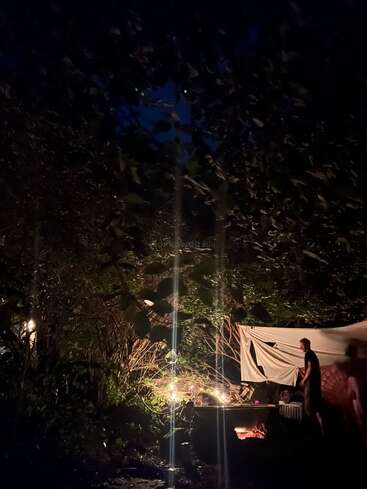 Uma cena ao ar livre pouco iluminada à noite, com um homem perto de uma fogueira brilhante. Árvores, folhas e uma lona ao redor criam uma atmosfera aconchegante e íntima.