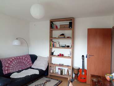 Esta sala de estar aconchegante tem um sofá preto com capas decorativas, uma estante de madeira, um violão, um tambor, uma lâmpada, um tapete e uma porta de madeira.