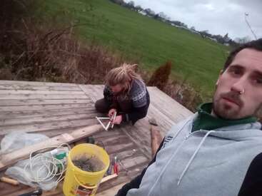 Two people are working outdoors on a wooden deck. One is taking a selfie, while the other is using tools. Supplies and a yellow bucket are nearby.