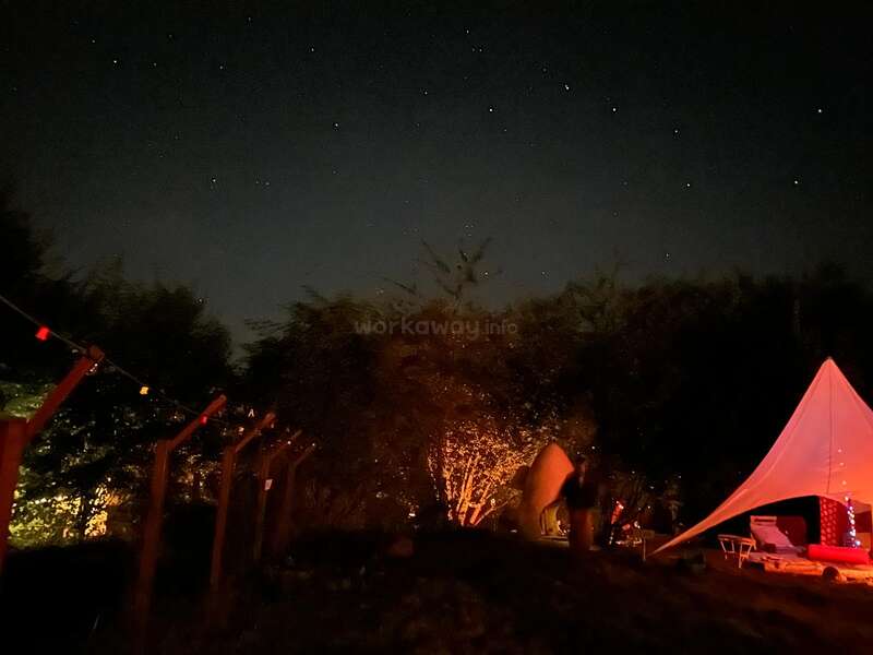 Um céu noturno estrelado acima de um acampamento com uma barraca brilhante, cadeiras aconchegantes, luzes de cordas e árvores iluminadas por uma fogueira quente, criando uma atmosfera tranquila.