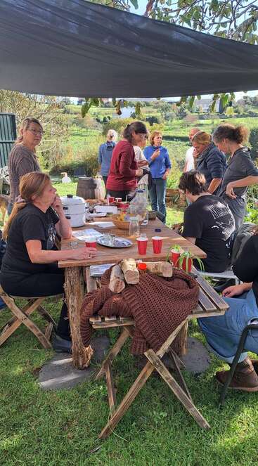 Eine Gruppe Leute trifft sich draußen unter einem Baldachin und genießt Essen, Getränke und Gespräche an einem rustikalen Holztisch in einer malerischen, grünen Landschaft.