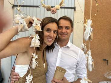 The image depicts a man and woman posing together, surrounded by decorative items, with the woman holding a string of small bags and the man holding a paper cup.