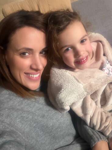 A woman and a young girl smile together, cozy and warm. They are snuggled up on a couch, sharing a happy, close, and loving moment.