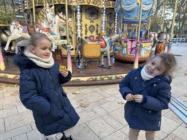Two smiling girls in warm coats stand in front of a colorful carousel. They appear happy, holding tickets, ready for a fun ride on a chilly day.