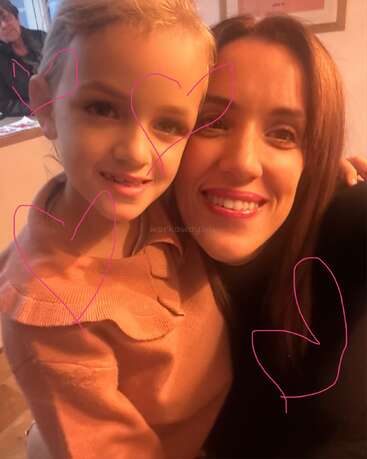 A woman and a young girl smiling close together, with pink hearts drawn over the photo. Warm lighting, cozy atmosphere, and cheerful expressions. Another person in the background.