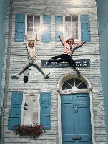 Two children appear to be climbing and hanging on a sideways house facade, creating a playful and whimsical optical illusion with blue shutters and a french street sign.