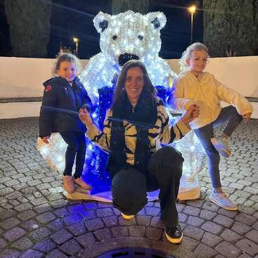 Eine Frau und zwei junge Mädchen posieren glücklich vor einer großen, beleuchteten Eisbärendekoration bei Nacht, umgeben von festlichen Lichtern und Outdoor-Kulissen.