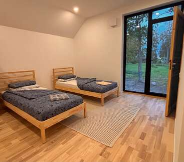 Essa imagem mostra um quarto aconchegante com duas camas de solteiro de madeira, roupa de cama azul, um tapete listrado, piso de madeira e janelas grandes com vista para uma paisagem verde ao ar livre.