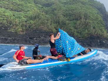 Four young men on a large paddleboard ride choppy ocean waves near a lush, green cliff. One holds a blue checkered blanket like a sail.