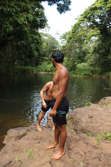 Two men in swim trunks stand on rocky riverbank surrounded by lush green forest, preparing to swim or jump into the calm, clear water on a sunny day.
