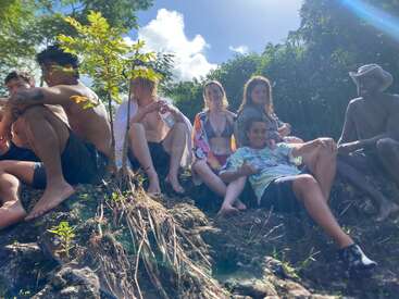 A group of friends relaxes together on a grassy riverside hill, enjoying the sunshine and nature, surrounded by greenery, wearing summer clothes and swimsuits.