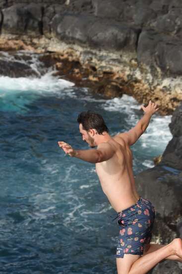 A man in floral swim trunks is mid-air, jumping off dark rocky cliffs into the ocean below. The waves crash against the jagged shore. Adventure awaits.