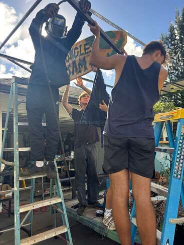 Three people are working together outdoors, assembling a metal frame using ladders. One person wears welding gear, while the others assist, holding a colorful “Jungle Jam” sign.