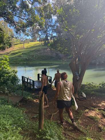 Four people approach a wooden dock by a serene river, surrounded by lush greenery and trees, enjoying a sunny day in nature and relaxation.