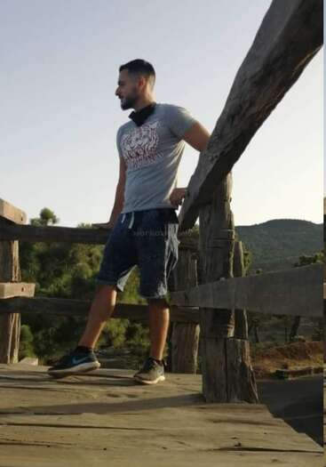 Das Bild zeigt eine Person, die vor einem Gewässer steht, mit einer Bergkette im Hintergrund und einem klaren Himmel darüber. Die Person trägt einen Rucksack.