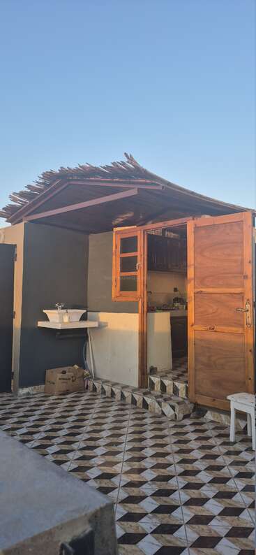 Ein kleines Gebäude mit einer Holztür, einem Waschbecken im Freien, einem gemusterten Fliesenboden, einem rustikalen Holzdach und einer Kiste auf dem Boden. Der Himmel ist klar, das Sonnenlicht erhellt die Szene.