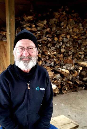 A smiling elderly man with a white beard and glasses, wearing a black beanie and jacket, sits indoors in front of a large stacked firewood pile.