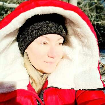 A woman wearing a thick red winter coat with a furry hood and a black knit hat enjoys bright sunshine outdoors, surrounded by a wintery natural setting.