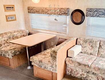 This image shows a cozy RV or camper dinette area with floral upholstered seating, a rectangular table, decorative wall art, a mirror, and window blinds.