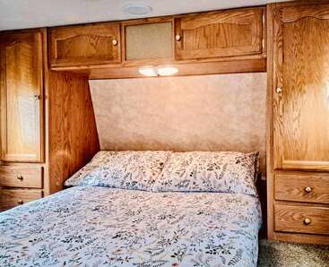 A cozy bedroom with a floral-patterned bedspread, surrounded by warm wooden cabinets and drawers. Soft lighting above the bed creates a welcoming, comfortable atmosphere.