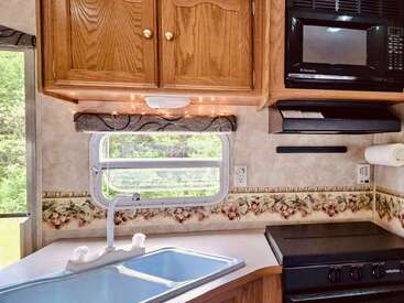 This image shows a cozy RV kitchen with wooden cabinets, a small sink, stove, microwave, paper towel holder, floral wallpaper, and a window overlooking greenery outside.
