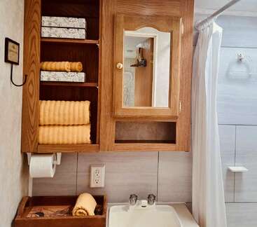 This image shows a tidy bathroom with a wooden cabinet, mirror, yellow towels, tissue boxes, a white sink, shower curtain, and a towel hanging hook.