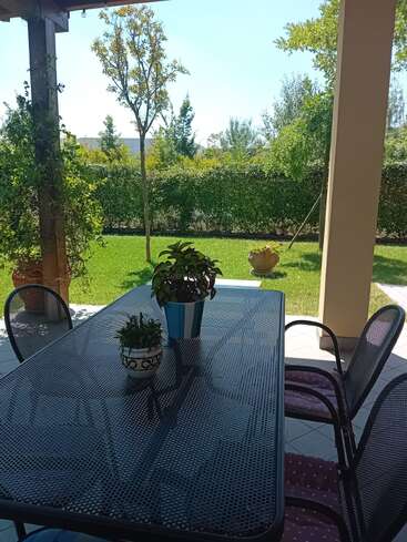 Um pátio ensolarado com uma mesa de metal preta e quatro cadeiras. Dois vasos de plantas estão sobre a mesa. Um jardim exuberante, árvores e vegetação cercam o espaço.