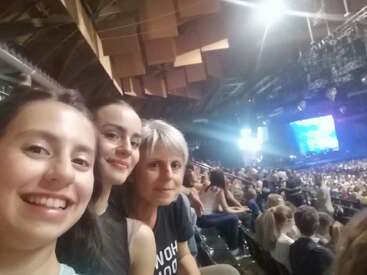 Tres personas sonrientes se toman un selfie en un concierto o evento en interior. El fondo muestra una gran multitud, luces brillantes y un escenario con pantallas de colores.