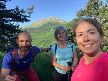 Tres personas disfrutan de un soleado día de senderismo en la montaña. Están sonriendo, rodeados de frondosos árboles verdes, con una vista panorámica de la montaña detrás de ellos.