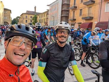 Dos ciclistas en primer plano sonríen y posan para un selfie, rodeados de un gran grupo de corredores en un bullicioso entorno urbano, listos para una carrera.