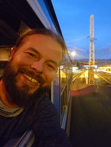 Ein bärtiger Mann lehnt sich in der Abenddämmerung freudig lächelnd aus dem Fenster eines Zuges. Die Lichter des Bahnhofs beleuchten die Gleise und den Bahnsteig und schaffen eine warme, einladende Atmosphäre.