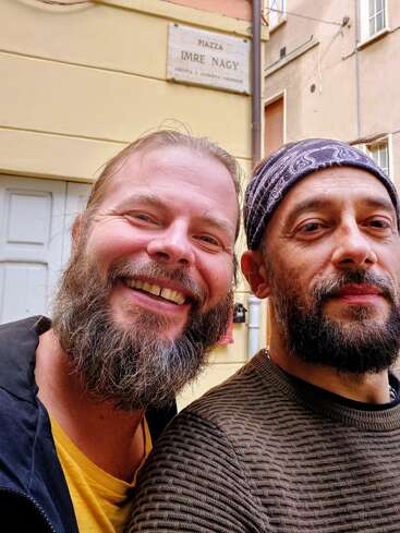 Zwei bärtige Männer posieren eng beieinander auf einer Straße, der eine lächelt breit, der andere ist neutral. Hinter ihnen steht ein Schild mit der Aufschrift "Piazza Imre Nagy".