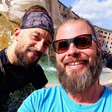 Two bearded men smiling for a selfie, one wearing sunglasses, the other with a bandana. They're outdoors, in front of a beautiful fountain on a sunny day.