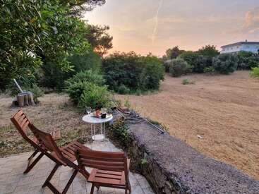 Dos sillas de madera y una mesita con bebidas y aperitivos dan a un tranquilo campo al atardecer, rodeadas de árboles, arbustos y una casa en la lejanía.
