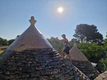 Una persona vestida con ropa deportiva está limpiando el tejado cónico de piedra de una casa tradicional de tipo trullo con una hidrolimpiadora, bajo un sol radiante y un cielo despejado.