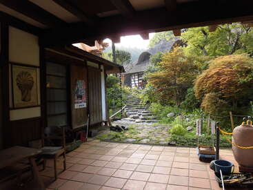 L'image représente une sereine maison de style japonais avec un porche carrelé, entourée d'une végétation luxuriante et d'un sentier en pierre menant à un bâtiment japonais traditionnel à l'arrière-plan.